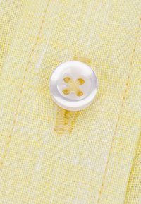 Żółty materiał z jednym białym guzikiem, mającym cztery dziurki i widoczną nitkę. Tekstura jest tkana i lekko chropowata.
