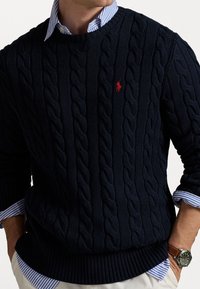 Pull en maille torsadée bleu marine avec ourlet côtelé, arborant un petit logo poney rouge. Porté par-dessus une chemise à col rayée bleue et blanche.