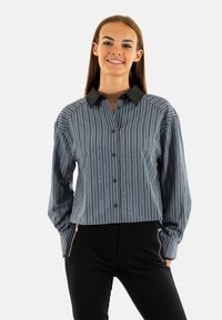 Blusa a rayas azul y blanca con cuello negro, cierre de botones en la parte delantera y mangas largas. Presenta líneas verticales y patrones sutiles.