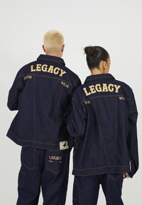 Tmavomodré džínové bundy s pozláteným vyšívaným textom "LEGACY" a "DENIM WEAR" na chrbte, s kontrastným šitím a uvoľneným strihom.