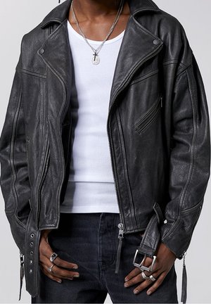 Person trägt eine schwarze Leder-Bikerjacke über einem weißen, gerippten Tanktop, dunkle Jeans, eine silberne Kreuzkette und mehrere silberne Ringe.