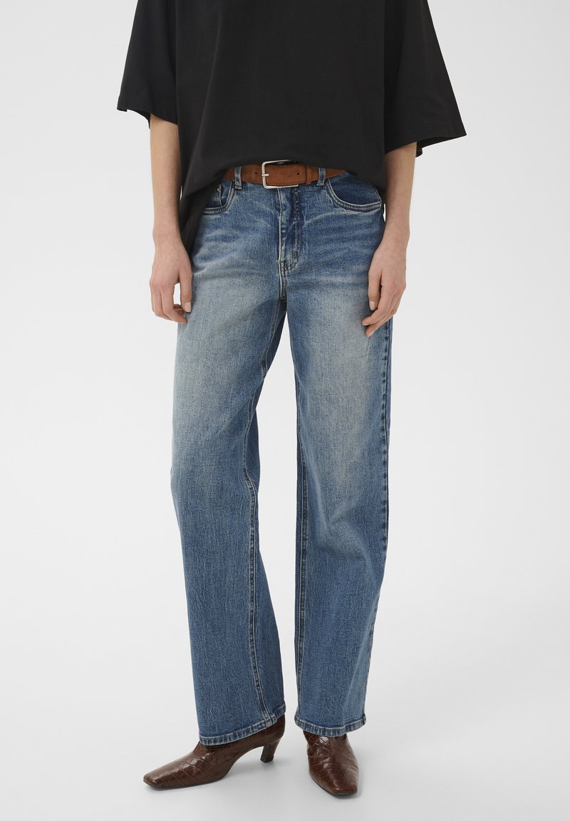 Personne portant un jean bleu délavé droit, une ceinture marron, un t-shirt noir oversize et des bottes marron à talons, debout devant un fond uni.