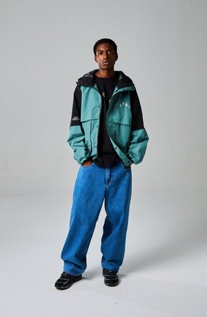 Rompevientos verde azulado y negro con múltiples bolsillos, combinado con una camiseta negra y jeans azules holgados, y zapatillas negras, posando frente a un fondo gris.