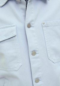 Svijetloplavi traper jakna s srebrnim kopčama, dva prednja džepa i teksturiranom tkaninom. Ima klasičan ovratnik i šivane detalje.