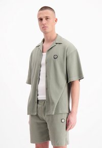 Khaki kortemouwige overhemd met knopen van een gestructureerde stof, gecombineerd met bijpassende shorts en een witte tanktop. Bevat een logo-embleem op elk kledingstuk.
