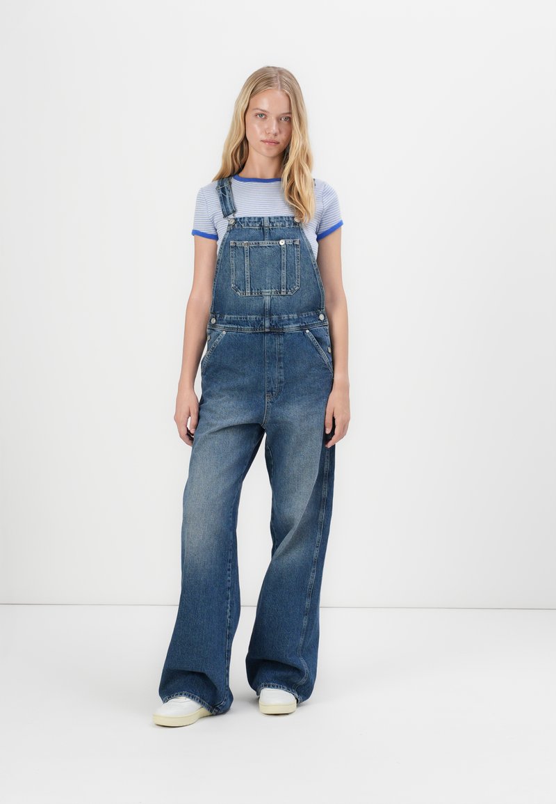 Jeanslatzhosen mit weitem Bein, ausgestattet mit verstellbaren Trägern, vorderen Taschen und einem blauen gestreifen Hemd darunter.