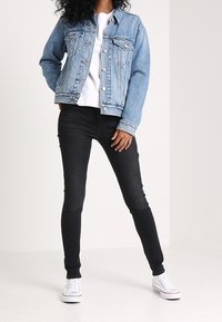 Modrá denimová bunda s knoflíkovým zapínáním, dvěmi náprsními kapsami, bílá mikina pod ní, spárovaná s černými skinny jeans a bílými teniskami.