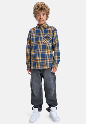 Chłopiec z kręconymi włosami, ubrany w niebieską i żółtą koszulę w kratę, luźne ciemne dżinsy oraz białe trampki na wysokiej podeszwie, stojący na białym tle.