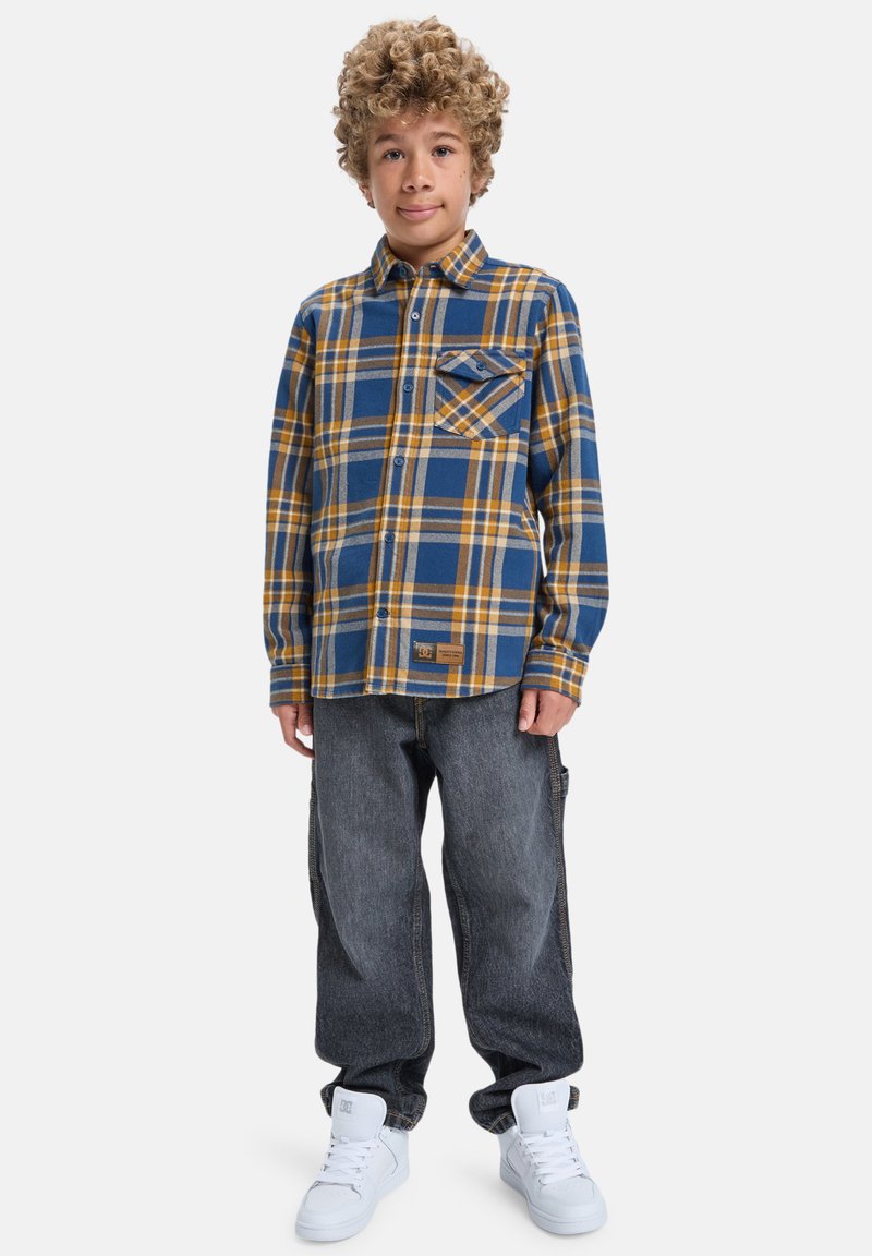 Chlapec s kučeravými vlasmi, oblečený v modro-žltom kockovanom tričku, vo voľných tmavých džínsoch a bielych vysokých teniskách, stojí na bielom pozadí.