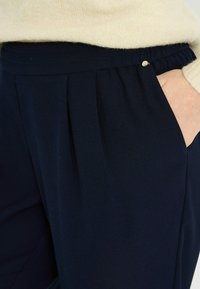 Pantalones plisados de color azul marino con cintura elástica, que cuentan con bolsillos inclinados y un pequeño detalle metálico cerca de la cintura. Textura de tela suave.