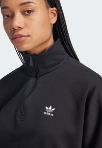 Mustard värvi tõmblukuga dressipluus, mis on valmistatud puuvillasegusest materjalist, kõrge kaelusega, tekstuurse pinnaga ja väikese valge Adidas'i logoga rinnal.