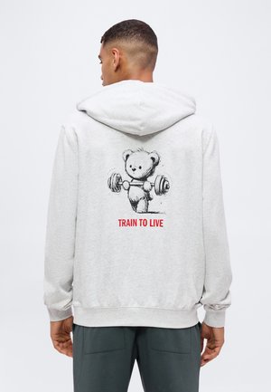 Homme portant un sweat à capuche gris clair avec un ours en dessin animé soulevant une barre et le texte rouge "TRAIN TO LIVE" dans le dos.