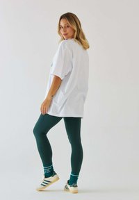 Biała oversize'owa koszulka, zielone legginsy, turkusowe skarpetki i kremowe sneakersy z ciemnymi akcentami. Ubranie ma luźny krój i casualowy styl.