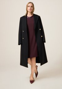 Czarny wełniany płaszcz z złotymi guzikami, noszony na bordowej sukience. Modelka ma na sobie bordowe lakierowane szpilki. Prosta, elegancka sylwetka.