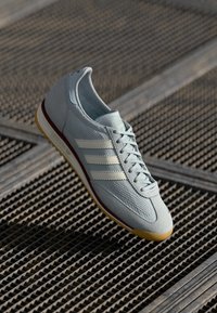 Sapatilha atlética azul-claro com parte superior em malha, três riscas brancas, sola de borracha texturizada e detalhes em castanho. Apresenta atacadores e colarinho acolchoado.