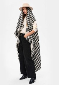 Czarna i biała kocykowa narzuta w kratę z frędzlami, noszona na jasnym swetrze i czarnych szerokich spodniach, zestawiona z beżowym kapeluszem.