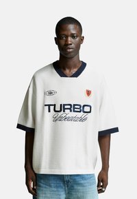 Young man wearing a white oversized T-shirt with navy trim and "TURBO Undetectable" printed on front, paired with light blue jeans.