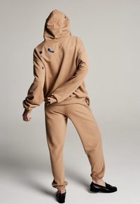 Hoodie surdimensionné marron clair avec capuche et poches, présentant des accents bleus et des motifs irréguliers. Associé à un pantalon de jogging assorti.