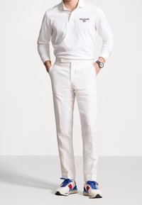 Vit bomull långärmad polo med "POLO SPORT"-logotyp, tillsammans med vita linbyxor och flerfärgade sneakers.