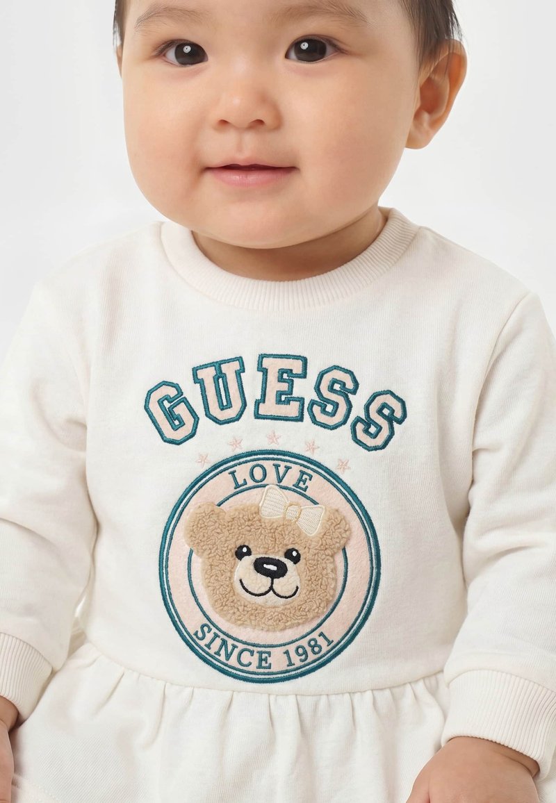 Bébé portant un sweat-shirt blanc avec un motif d'ours en peluche et le texte "GUESS LOVE SINCE 1981" sur le devant.