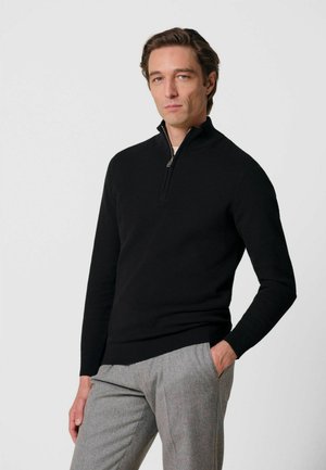 Hombre con un suéter negro con cremallera hasta el pecho y pantalones gris claro, de pie con una mano en el bolsillo contra un fondo claro y simple.