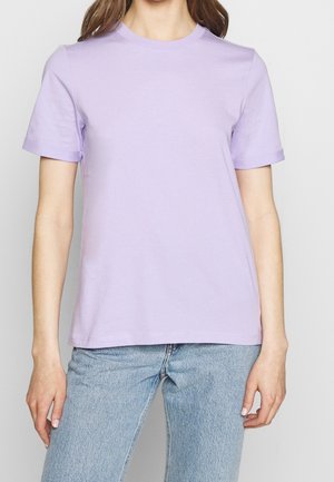 Femme portant un t-shirt uni lavande à manches courtes et col rond, ainsi qu'un jean en denim bleu clair, debout avec les bras détendus le long du corps.