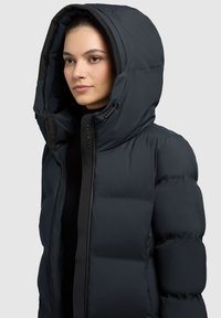 Puffer bunda v tmavě námořnické barvě s velkou kapucí, se zipovým zapínáním a hladkým, matným povrchem. Doplněna odolnými kovovými detaily.