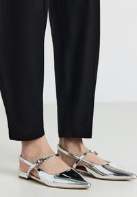 Chaussures plates argentées avec un bout pointu, une sangle au talon et une boucle à la cheville. Fini métallique lisse et petit talon. Assorties à un pantalon noir.