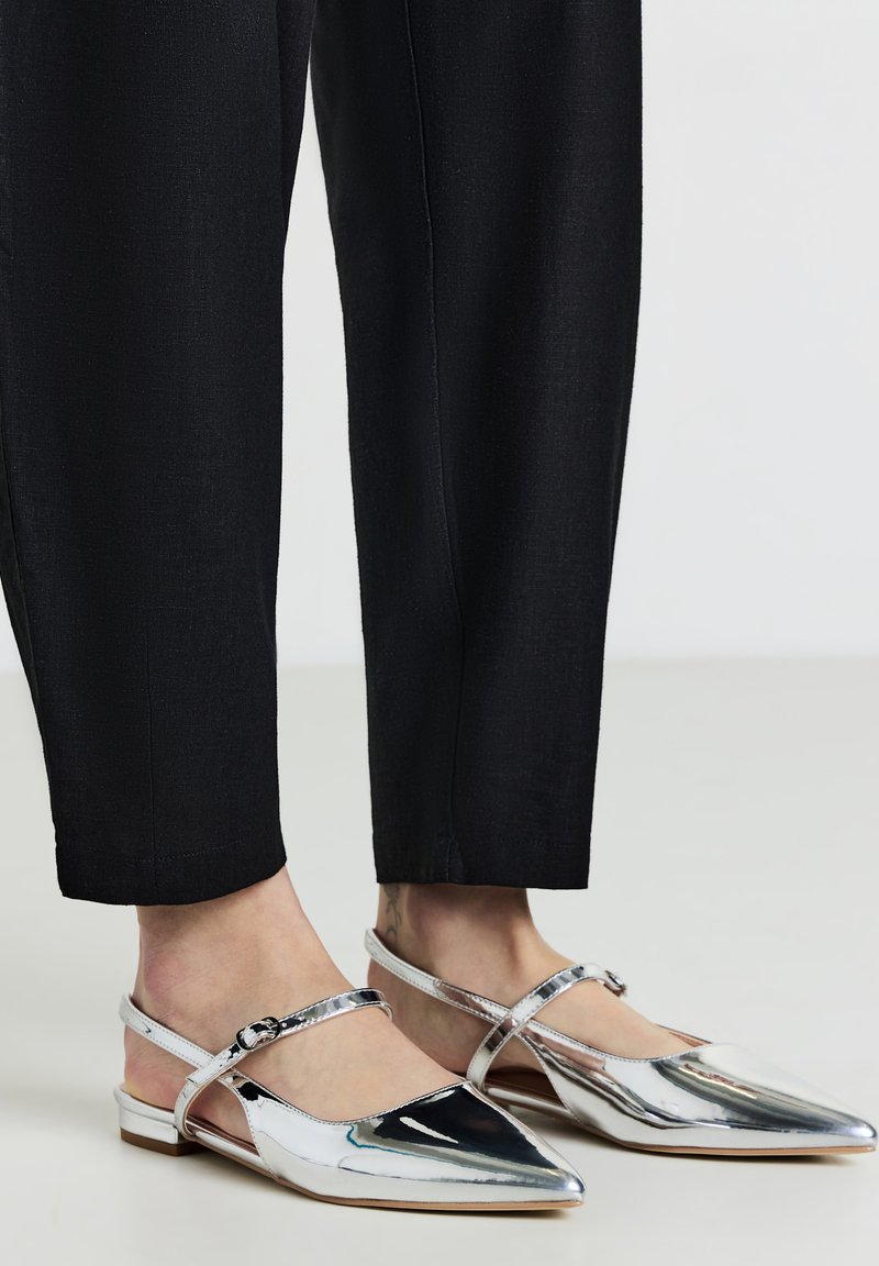 Chaussures plates argentées avec un bout pointu, une sangle au talon et une boucle à la cheville. Fini métallique lisse et petit talon. Assorties à un pantalon noir.