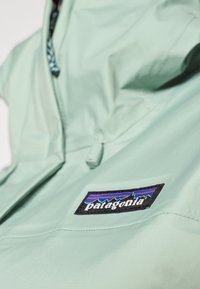 Gaiši zaļš Patagonia jaka ar rāvējslēdzēju priekšpusē, regulējamu apkakli un melnu logotipa uzlīmi ar krāsainiem kalnu grafikiem.