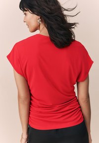 Červený top s krátkými rukávy, volného střihu a na zádech se shromážděnými detaily, vyrobený z měkké, hladké látky, v kombinaci s černou sukní.