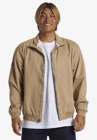 Light brown zip-up jacket with a stand collar, elastic cuffs, and logo on the sleeve. Worn over a white T-shirt, paired with dark jeans.