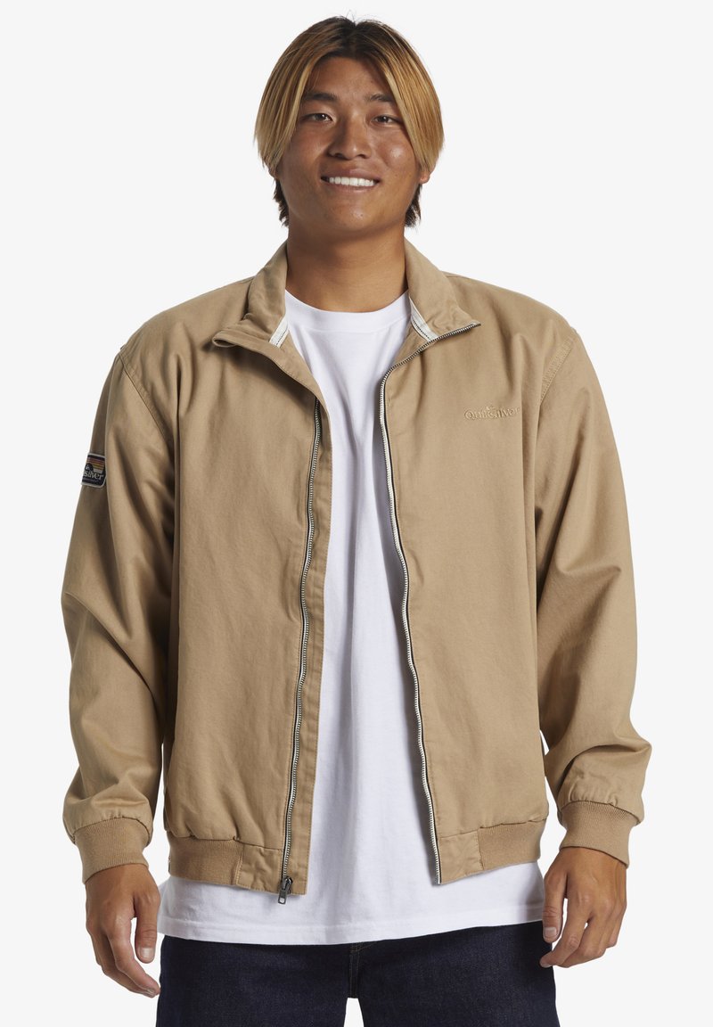 Light brown zip-up jacket with a stand collar, elastic cuffs, and logo on the sleeve. Worn over a white T-shirt, paired with dark jeans.