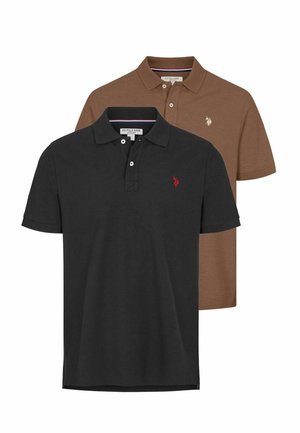 Homme debout portant un polo marron à manches courtes, un pantalon bleu foncé et des baskets blanches sur un fond clair uni.