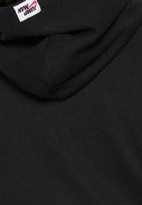 Tessuto di felpa nera con un'etichetta rettangolare bianca sul colletto, caratterizzata da un testo rosso e nero con un design a sottovento.