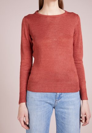 Femme portant un pull en maille ajusté à manches longues couleur rouille et un jean bleu clair sur un fond uni.