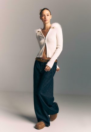 Young woman in a white zip-up sweater with fur trim, wide-leg dark jeans, brown belt, and brown platform shoes, standing against a neutral background.