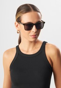 Rektangulära solglasögon med mörkt tonade linser och en matt olivgrön båge. Bäraren har på sig en strukturig svart linne och guldörhängen i form av ringar.