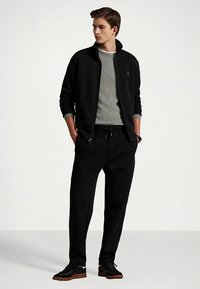 Czarna kurtka na zamek, pod spodem szary sweter, czarne spodnie dresowe i czarne sneakersy. Miękki materiał o luźnym kroju. Minimalistyczny design.