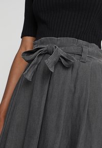 Šedá plisovaná sukně s látkovým páskem uvázaným do mašle v pase, s jemnou texturou a jednobarevným designem.