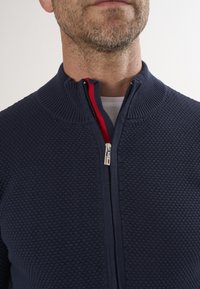 Tumesinine zip-up kampsun, millel on tekstureeritud mesilasvahatooted, soonikkoes krae ja eristuv punane aktsent tõmbluku sees.