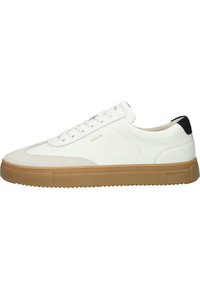 Des baskets blanches en cuir présentent une partie avant en daim texturé, une semelle en caoutchouc beige avec des nervures, et des accents de talon noirs avec un branding subtil.