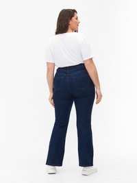 Jeans acampanados de color azul oscuro con costuras en contraste, que cuentan con bolsillos traseros y una cintura alta, combinados con una camiseta blanca lisa.
