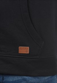 Musta collegepaita resorikähdyksillä, jossa on ruskea nahkainen merkki alhaalla edessä, jossa lukee "BLEND GLOBAL JEANMAKER".