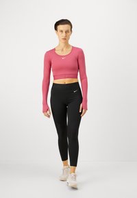 Rozā garām piedurknēm krekls ar apaļu dekoltē, kas sader ar melnajām leggings, kurām ir tekstūrēti paneļi un pieguloša forma. Gaisīgas sporta apavi.