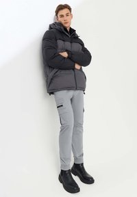 Giovane in piedi, indossa un giubbotto imbottito nero e grigio, pantaloni cargo grigi chiari e stivali neri robusti contro un muro bianco.