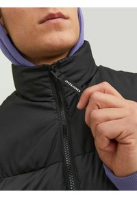 Black puffer jacket with a high collar and zipper; features a small label with "JACK&JONES," worn over a purple hoodie. Textured fabric.