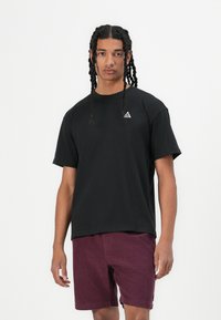 Schwarzes Baumwoll-T-Shirt mit Rundhalsausschnitt und kurzen Ärmeln, mit einem kleinen weißen dreieckigen Logo auf der Brust. Das Modell trägt burgunderfarbene Shorts.