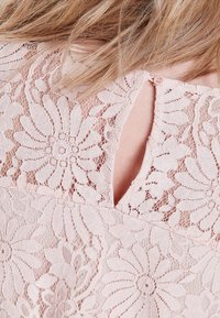 Spitzenoberteil in sanftem Rosa, mit floralen Mustern und einem Rückenschlitzdesign, das mit einem kleinen Knopf gesichert ist. Strukturierter, leichter Stoff.
