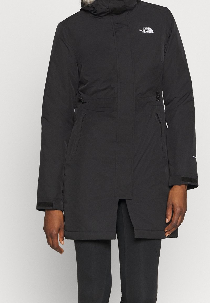 Parka noire isolante avec capuche bordée de fourrure, poches latérales zippées et logo "The North Face" sur la poitrine, portée par une personne debout.